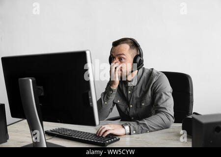 Confused or angry businessman or student in a shirt sitting against monitor of computer in headphones and speaking with somebody via internet. Working Stock Photo