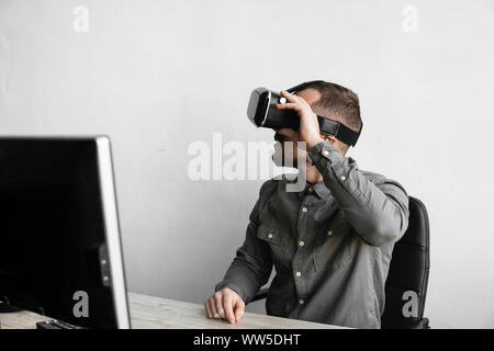 Young bearded man sitting against computer with virtual reality glasses. Modern technologies. The concept of future technology. Stock Photo
