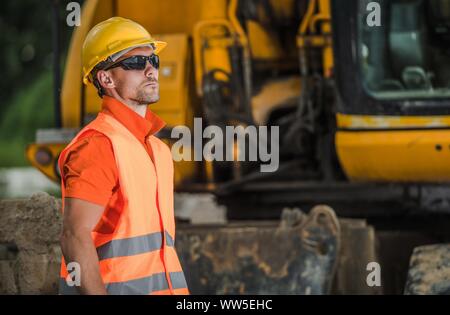 Construction Worker on Duty. Caucasian Men in His 30s Wearing Safety Accessories. Building Machinery in a Background. Industrial Zone. Stock Photo