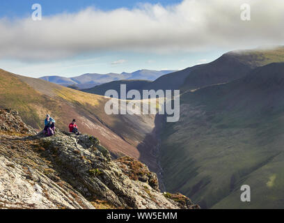 Two hikers sitting down on a rocky crag with distant views of Sand Hill and Crag Hill above Gasgale Gill in the English Lake District, UK. Stock Photo