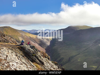 Two hikers sitting down on a rocky crag with distant views of Sand Hill and Crag Hill above Gasgale Gill in the English Lake District, UK. Stock Photo