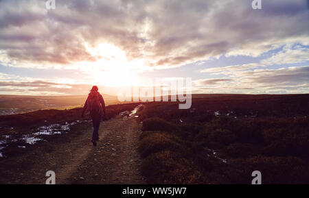 A hiker walking along with their dog on a dirt trail over open moorland at sunset at Edmunbyers in County Durham.