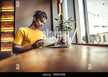 Stylish student with African hairstyle wearing casual clothes having doubtful look while looking at laptop computer not understanding new material try Stock Photo