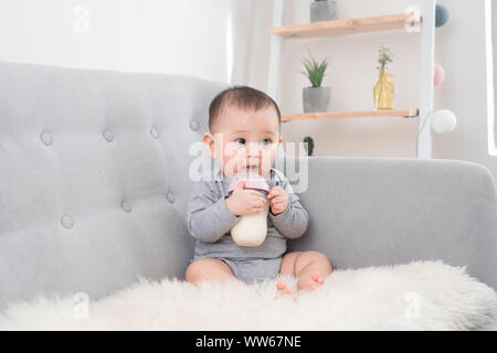 Little cute baby girl sitting in room on sofa drinking milk from bottle and smiling. Happy infant. Family people indoor Interior concepts. Childhood b Stock Photo