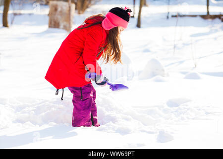 Beautiful little girl with long dark hear, wearing a red coat and playing shoveling the snow, on a cold winter day Stock Photo