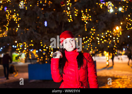 Close portrait of attractive young fashion model in bright red coat and wool mittens standing in front of city Christmas tree with lights on it. Winte Stock Photo