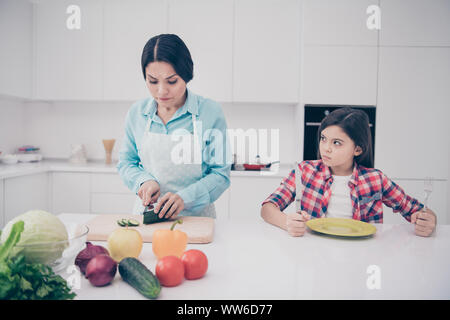 Portrait of two nice cute lovely attractive irritated annoyed bored tired girl demanding meal mature mom making lunch in light white kitchen interior Stock Photo