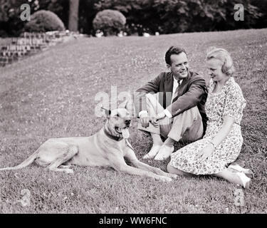 1930s SMILING COUPLE STYLISHLY CASUALLY DRESSED SITTING TOGETHER WITH GREAT DANE DOG ON WELL MAINTAINED HILLSIDE GARDEN LAWN - d34 HAR001 HARS ROMANCE BEAUTY SUBURBAN URBAN PET EXPRESSION OLD TIME FUTURE NOSTALGIA OLD FASHION 1 FACIAL STYLE COMMUNICATION BLOND SECURITY BALANCE SAFETY STRONG PLEASED JOY LIFESTYLE FEMALES MARRIED SPOUSE HUSBANDS HEALTHINESS HOME LIFE COPY SPACE FRIENDSHIP FULL-LENGTH LADIES PERSONS CARING MALES CONFIDENCE EXPRESSIONS B&W PARTNER SUCCESS SUIT AND TIE HAPPINESS MAMMALS WELLNESS CHEERFUL LEISURE PROTECTION STRENGTH CANINES EXTERIOR PRIDE ON SMILES POOCH CONNECTION Stock Photo