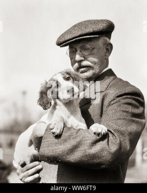 1920s IRISH RED AND WHITE SETTER PUPPY HELD IN ARMS BY SENIOR MAN WHITE MUSTACHE WIRE RIM GLASSES WEARING TWEED HAT AND COAT - d848 HAR001 HARS TWEED RURAL COPY SPACE HALF-LENGTH PERSONS INSPIRATION CARING MALES SENIOR MAN SENIOR ADULT B&W MUSTACHE TEMPTATION SUIT AND TIE HAPPINESS MAMMALS OLD AGE OLDSTERS OLDSTER MUSTACHES AND CANINES CHOICE EXCITEMENT HELD SETTER PRIDE BY AUTHORITY FACIAL HAIR ELDERS POOCH CONNECTION CONCEPTUAL STYLISH WOOLEN PERSONAL ATTACHMENT UPLAND WIRE RIM AFFECTION CANINE EMOTION GROWTH MAMMAL TOGETHERNESS BLACK AND WHITE CAUCASIAN ETHNICITY HAR001 IN ARMS Stock Photo