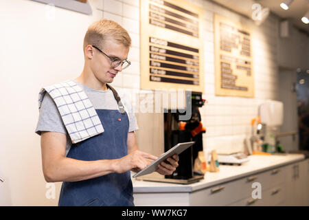 Young barista in workwear using digital tablet to enter new menu of drinks Stock Photo