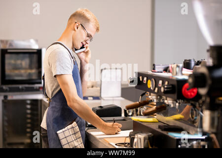 Young waiter or worker of cafeteria writing down orders of clients in notepad Stock Photo