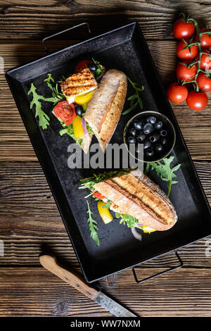 Wholemeal vegetarian baguette stuffed with grilled white cheese, arugula, tomatoes, black olives, bell pepper and red onion on wooden background Stock Photo