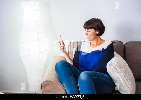 beautiful young woman at home sit down on the sofa speaking in live with friends with mobile phone technology. wifi friendship concept in bright house. Stock Photo