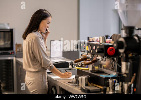 Female owner of cafeteria calling clients and making working notes on paper Stock Photo