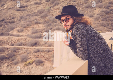 long hair beautiful young man drinking beer outdoor on the rooftop terrace with mountain and nature in background. vingtage retro filter. black hat and eyeglasses Stock Photo