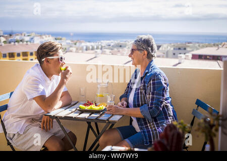happy leisure activity on the terrace rooftop having breakfast with smiles and happiness for grandmother and teenager family caucasian people. ocean and buildings view, outdoor coule together Stock Photo