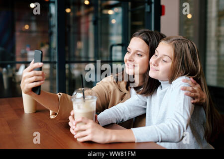 Affectionate mother and daughter with drinks looking at smartphone camera