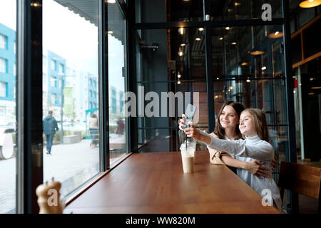 Happy girl stretching arms with smartphone while making selfie with mom in cafe Stock Photo
