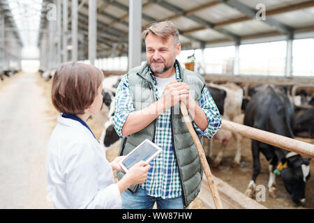 Happy farmer with hayfork looking at colleague with tablet during conversation Stock Photo