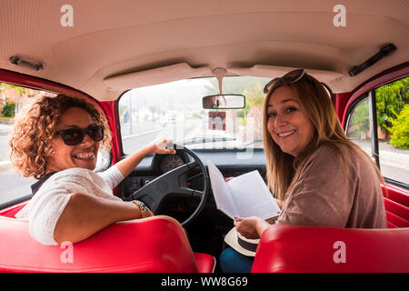 couple of caucasian middle age woman in outdoor leisure activity near and sitting on a red vintage car ready to travel, Stock Photo