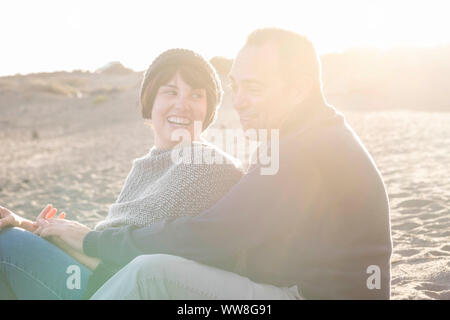 nice beautiful middle age 40 years old couple man and woman caucasian hugged and stay together on the beach sitting in the sand and enjoying a golden amazing sunset for nice leisure activity outdoor in the nature Stock Photo