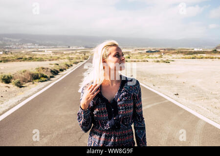 beautiful lonely blonde caucasian young girl walk in fashion dress in a long road in the middle of the desert, travel and independence freedom concept for smile people, sunlight and summer season Stock Photo