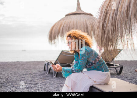 alternative office at the beach with ocean in background for a beautiful middle age woman working on her smartphone sitting on a seats under the umbrella sun, vacation lonely lady connecting with friends Stock Photo