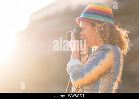 woman caucasian photographer with nice coloured hat taking pictures with old little camera, sunlight and sun flare in the background, bright summer concept image of nice people active taking memories Stock Photo