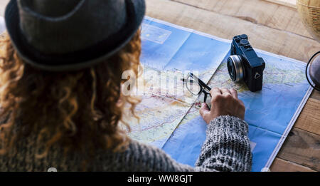 Hipster curly blonde female viewed from rear planning the new travel vacation wanderlust on the map, eyeglasses and camera on the wooden table with a globe worldwide sphere to choose and dream the new destination Stock Photo