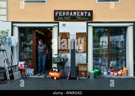 Exterior of an old hardware store with the shopkeeper on the threshold and goods displayed on the sidewalk in the city of Aosta, Aosta Valley, Italy Stock Photo