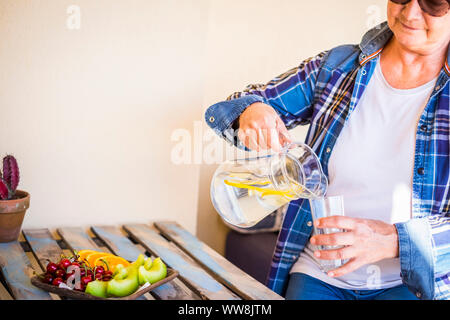 senior adult lady drink water and lemon detox drink to depurate body and stay healthy. nice lifestyle with fresh vegan and vegetarian food. morning break outdoor in the terrace at home Stock Photo