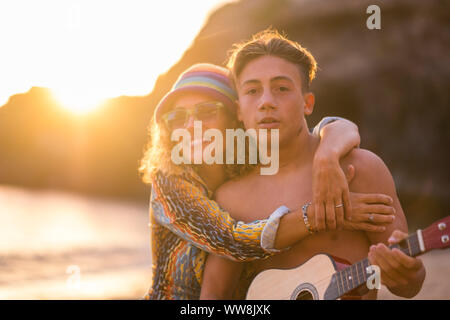 family leisure activity at the beach during a golden colored sunset in vacation and freedom lifestyle. mother and son together hugging and playing music with acoustic guitar. happiness concept Stock Photo