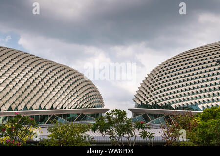 Singapore - March 20, 2019: Closeup of Double Esplanade Theatres domes under heavy cloudscape. Shot with entrance to both in center. Fronted by green Stock Photo