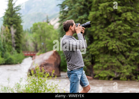 Man taking photos with camera in Redstone, Colorado during summer with large boulder and red bridge on Crystal river by trees Stock Photo