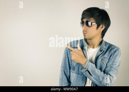 Young Asian Man in Jeans Jacket or Denim Jacket Wear Sunglasses and Pointing Finger. Denim or Jeans Jacket Men Fashion on Gray Background Stock Photo
