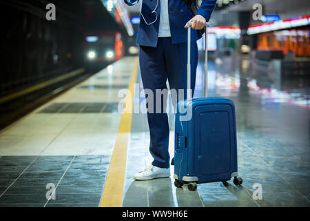Young stylish handsome man in suit with suitcase standing on metro station holding smart phone in hand, scrolling and texting, smiling and laughing. Stock Photo