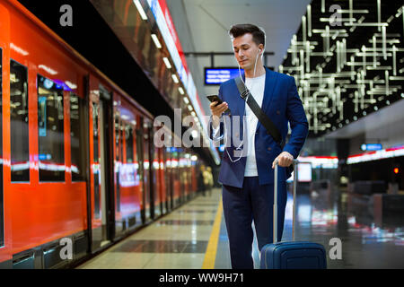 Young stylish handsome man in suit with suitcase standing on metro station holding smart phone in hand, scrolling and texting, smiling and laughing. Stock Photo