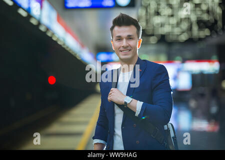 Young stylish handsome man in suit with suitcase standing on metro station holding smart phone in hand, scrolling and texting, smiling and laughing. Stock Photo