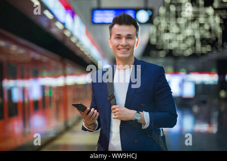 Young stylish handsome man in suit with suitcase standing on metro station holding smart phone in hand, scrolling and texting, smiling and laughing. Stock Photo