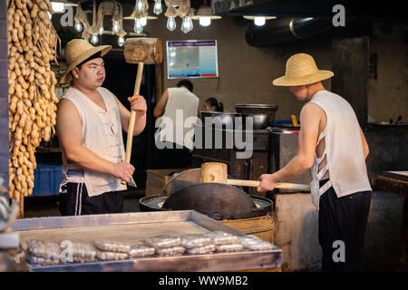 Xian, China -  July 2019 : Two men hitting hard with big wooden hammers to crack grain which will be used in making of sweet snack for sale on the str Stock Photo