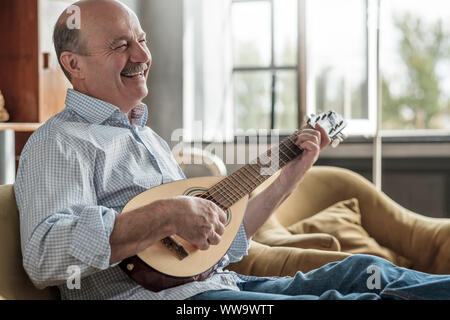Senior hispanic man playing guitar singing and laughing. Old man hold guitar sitting at the living room Stock Photo