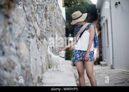 Naxos, Greece - June 28, 2018: A woman in flowered shorts pets a local cat in the village of Koronos on the island of Naxos, a popular destination. Stock Photo