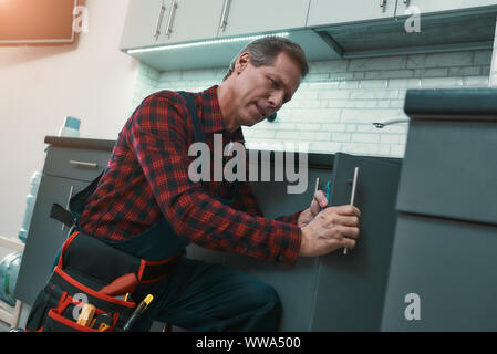 Portrait of mature man wearing tool kit standing in the kitchen, holding a drill and fixing the cabinet door. Horizontal shot Stock Photo