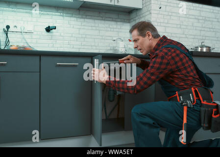 Portrait of mature man wearing tool kit standing in the kitchen, holding a screwdriver and fixing the cabinet door. Horizontal shot. Side view Stock Photo