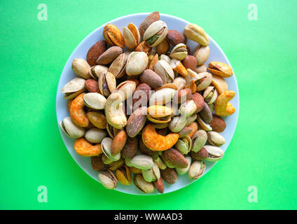 Mixed nuts on white plate isolated on green background. Healthy snack and food. Salted and spicy pistachios, cashew and almond. Top view. Stock Photo
