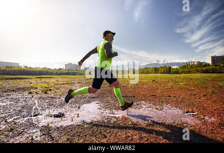 Man with grey beard running on the dirty trail with water splashes at mountain background in the morning. Healthy lifestyle concept Stock Photo