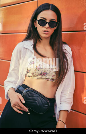 Portrait of girl in dark sunglasses posing in city against orange building. Dressed in top with floral print, white shirt, black trousers, waist bag. Stock Photo