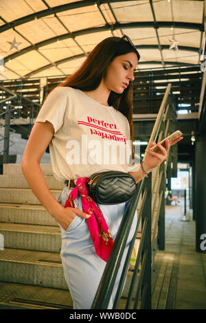 Portrait of a girl posing near a shopping mall, holding smartphone. Dressed in white t-shirt, blue trousers, black waist bag. Stock Photo