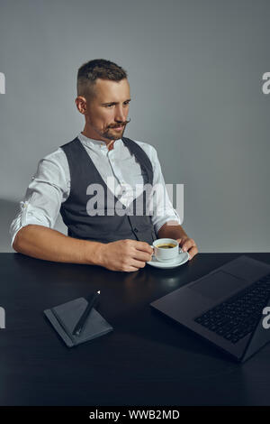 Bearded businessman with stylish mustache, dressed in a classic suit is looking at laptop while sitting at table in office, gray background. Stock Photo