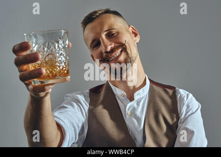 Man with stylish mustache, dressed in classic brown vest, white shirt is sitting at the table, enjoying whiskey. Grey background, close-up shot. Stock Photo